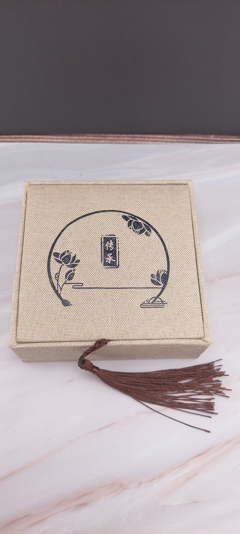 A beige Chinese-style jewelry box with a decorative pattern on the lid, a tassel hanging from the handle, and a white marble background.