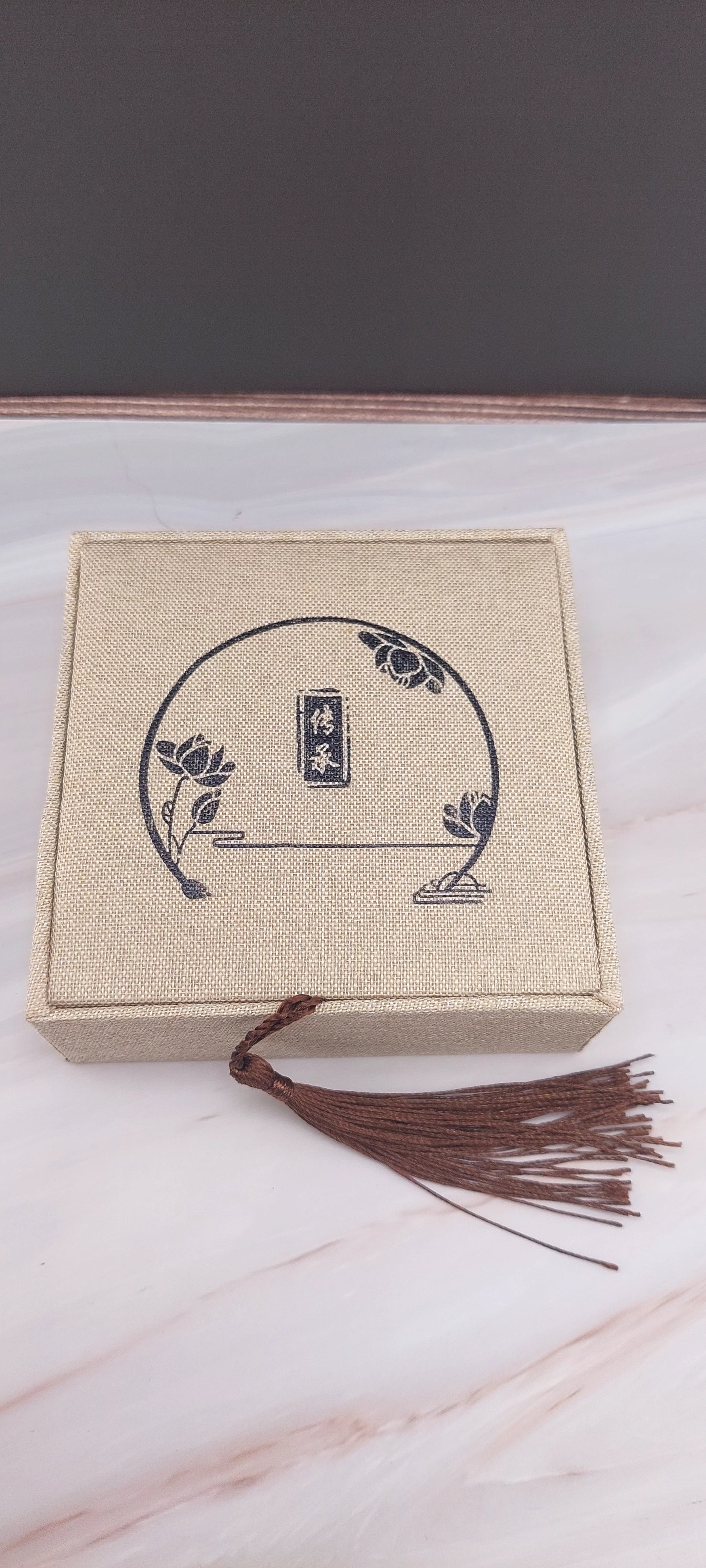 A beige Chinese-style jewelry box with a decorative pattern on the lid, a tassel hanging from the handle, and a white marble background.