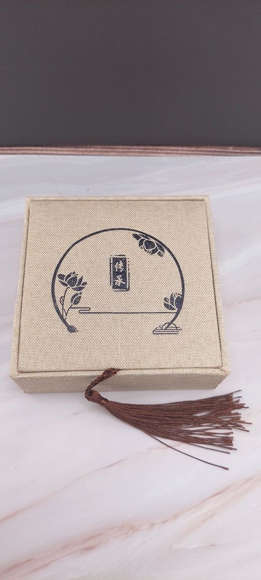A beige Chinese-style jewelry box with a decorative pattern on the lid, a tassel hanging from the handle, and a white marble background.