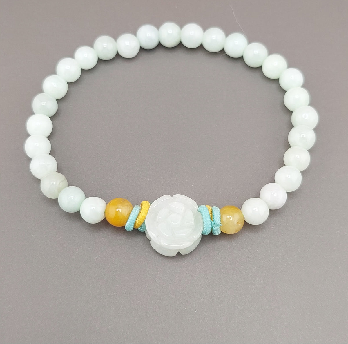 A bracelet made of round blue water jade stones with gold accents and a blue fabric tie at the clasp, featuring a small circular silver charm.