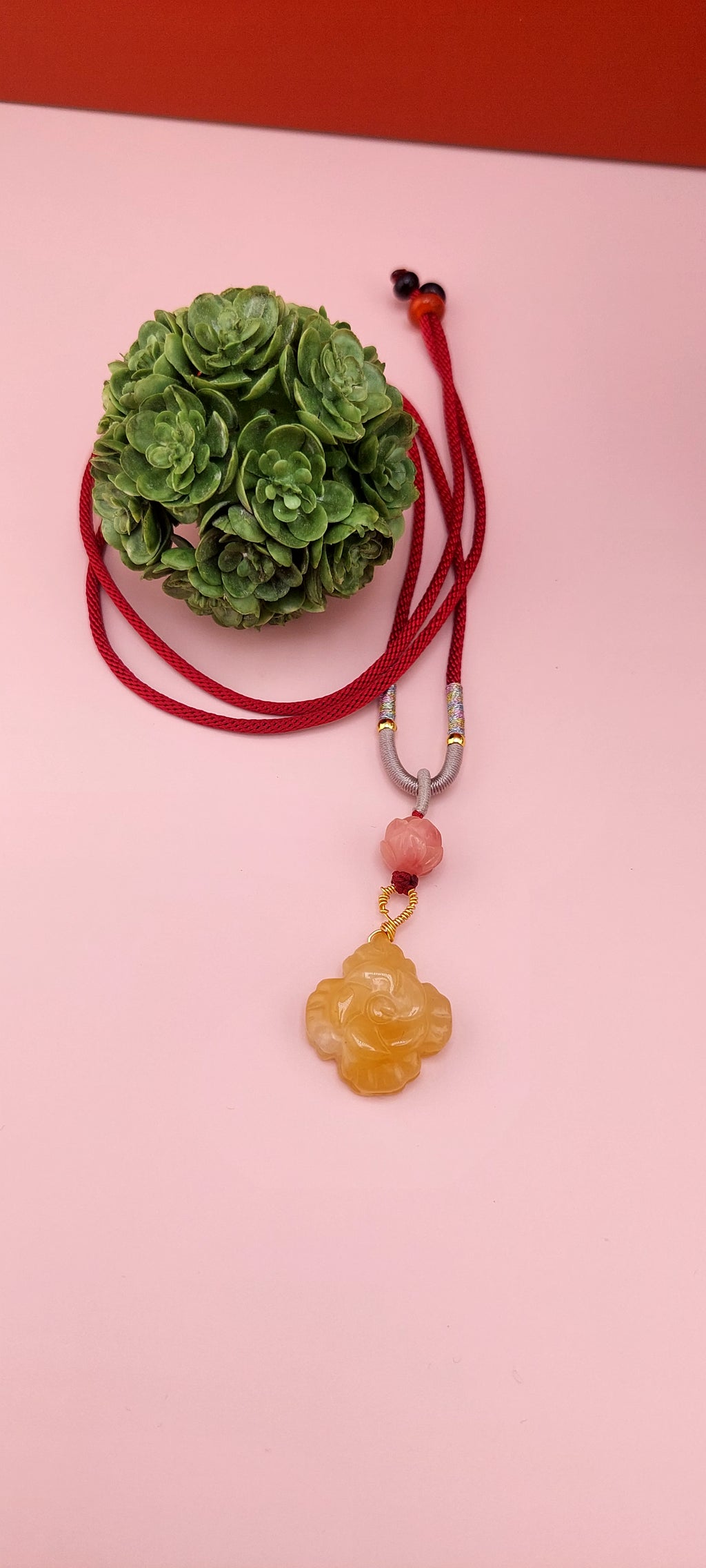 A colorful agate flower necklace with a red cord and a floral pattern on the agate stone, displayed against a pink background with a decorative green item in the background.