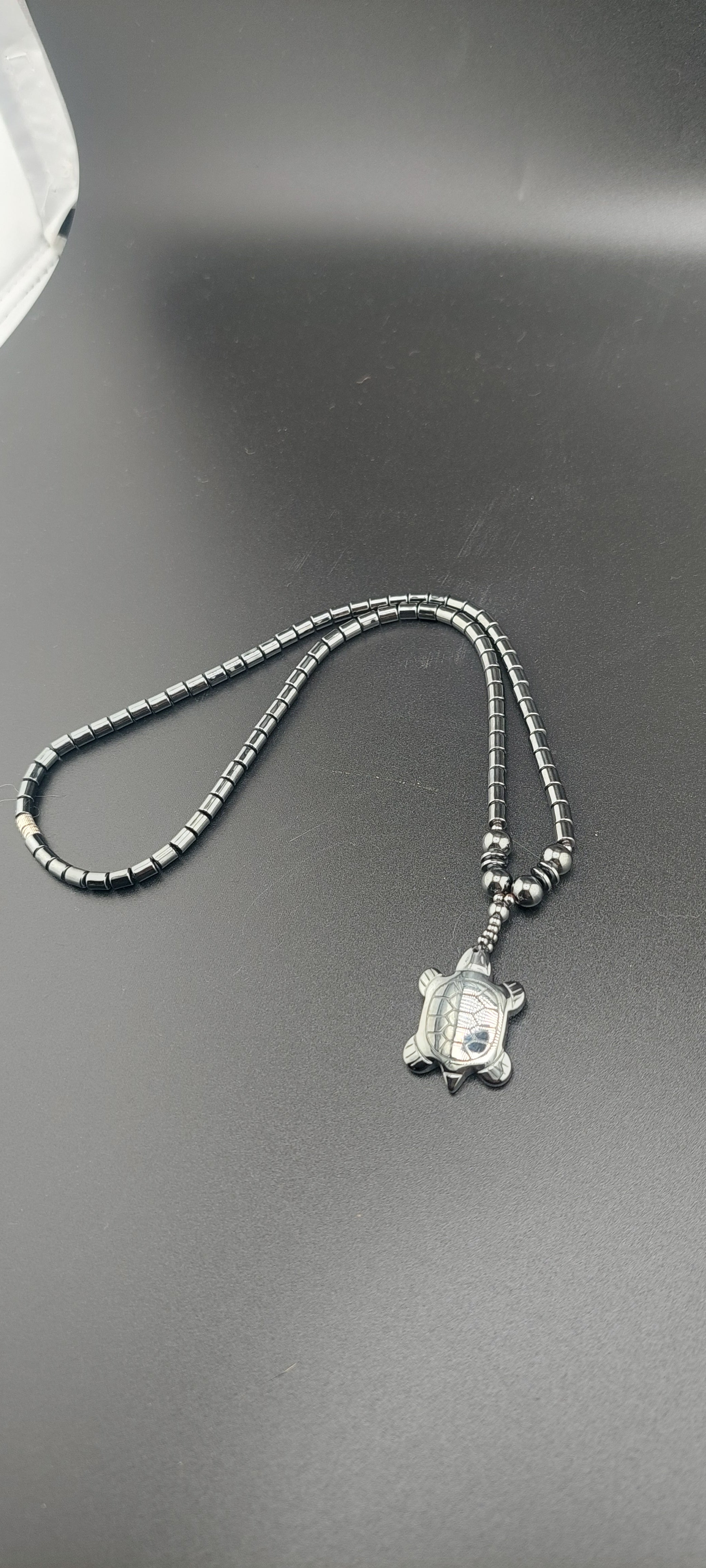A silver-colored hematite turtle necklace on a chain, displayed against a grey background.
