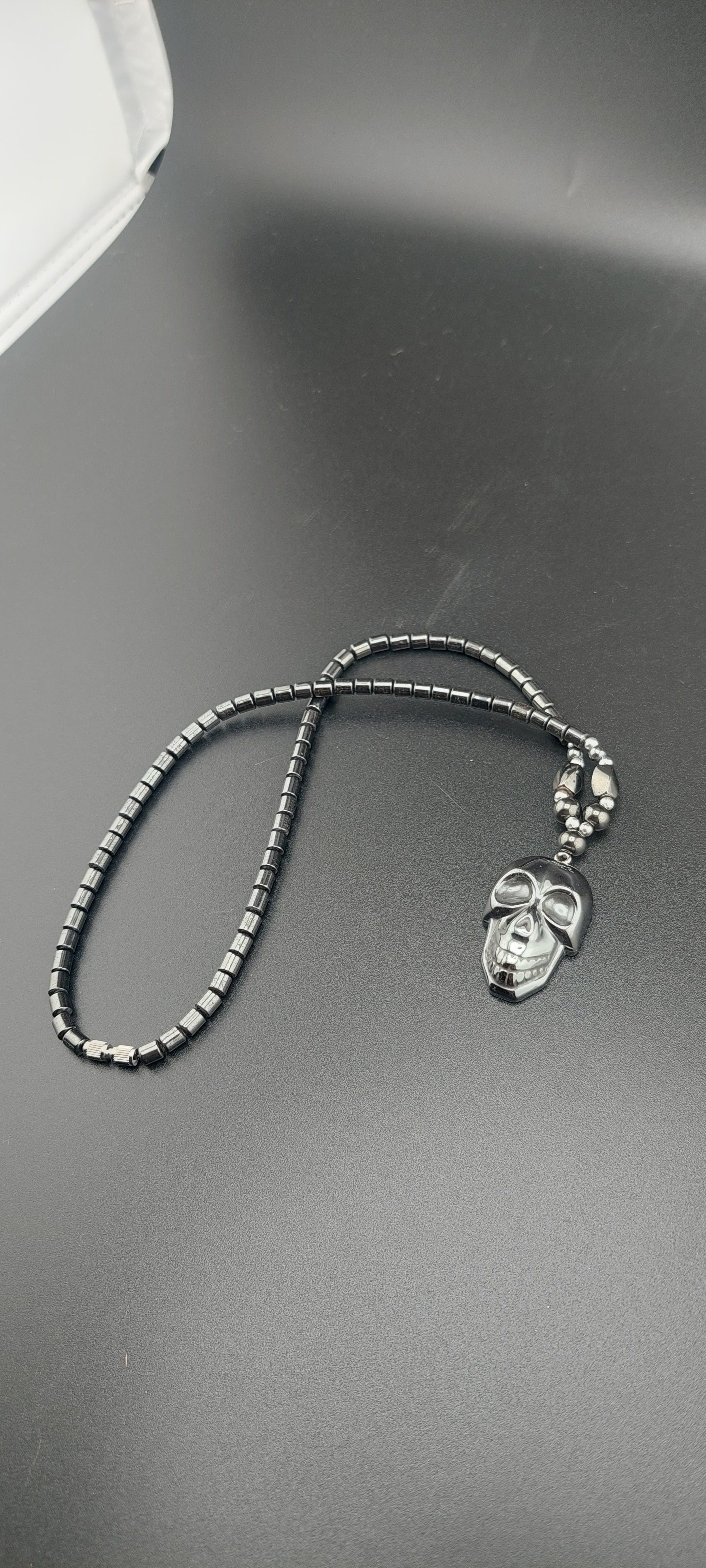 A black men's skull necklace with a hematite stone chain and a metallic skull pendant, displayed on a mannequin.