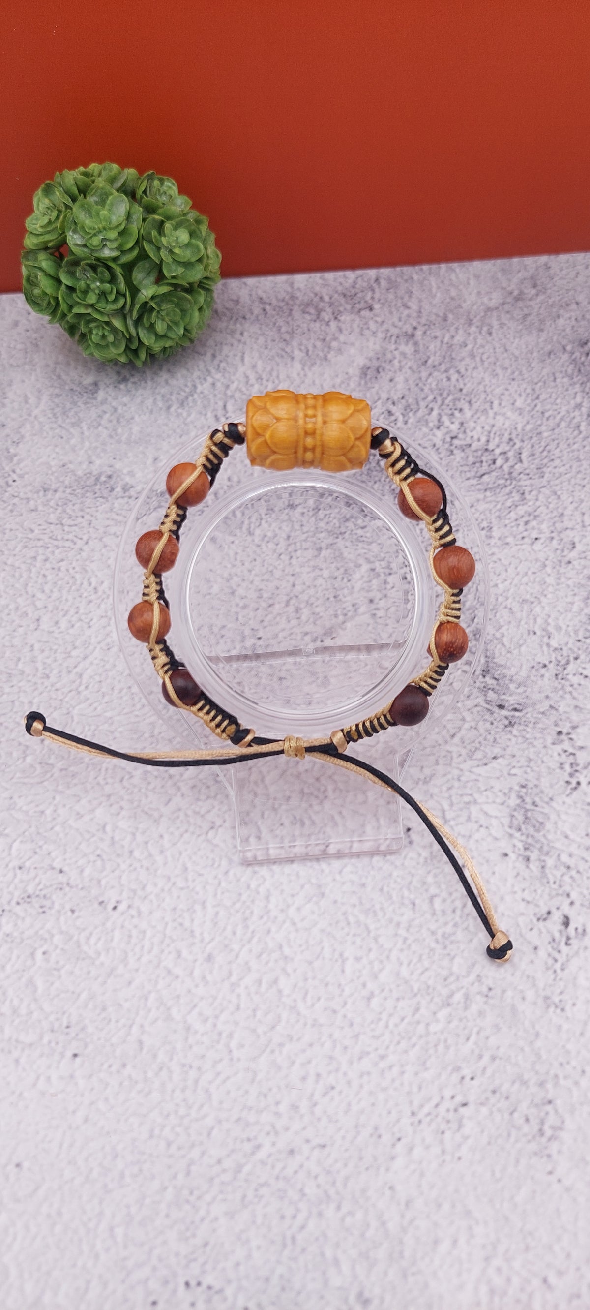 An adjustable nylon rope bracelet with macrame details and double wooden lotus bow design, displayed against a red background with a decorative ball to the left.
