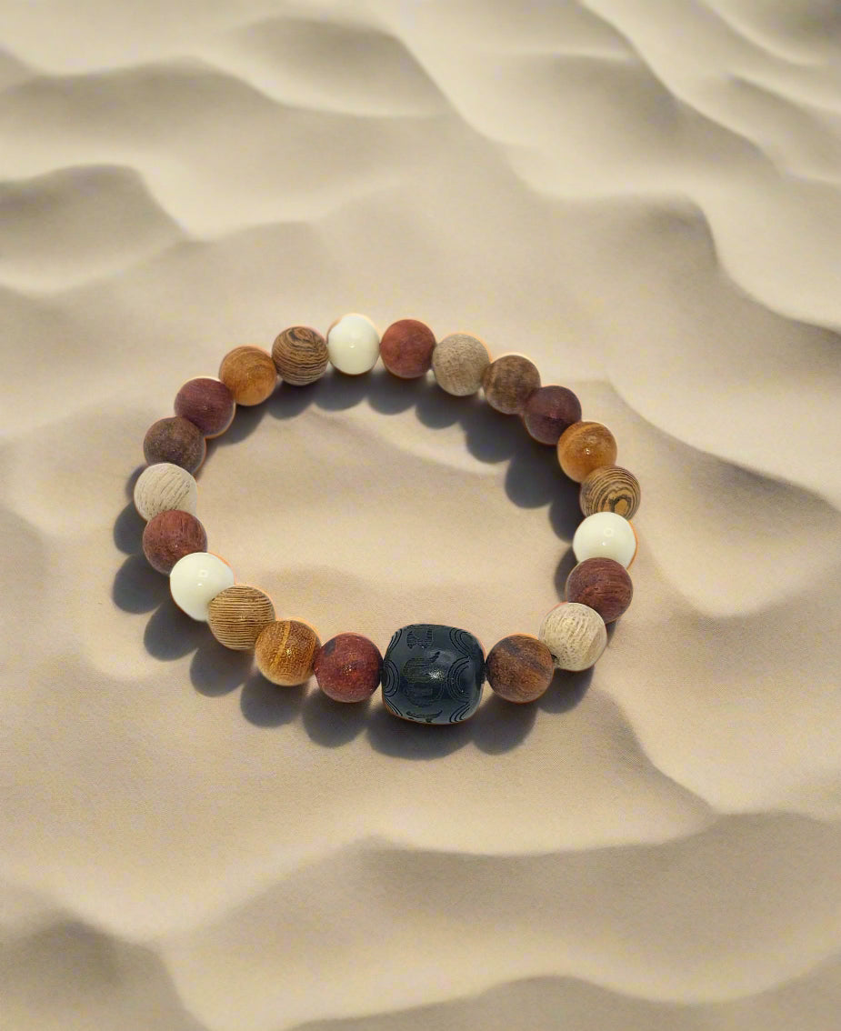 A sandle wood bracelet with a mix of brown, beige, white, and a single black bead, displayed on a sandy surface.